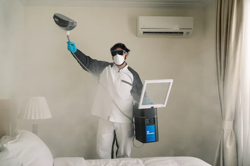 Technician in mask and gloves fogging near an open AC return grille in an empty apartment bedroom
